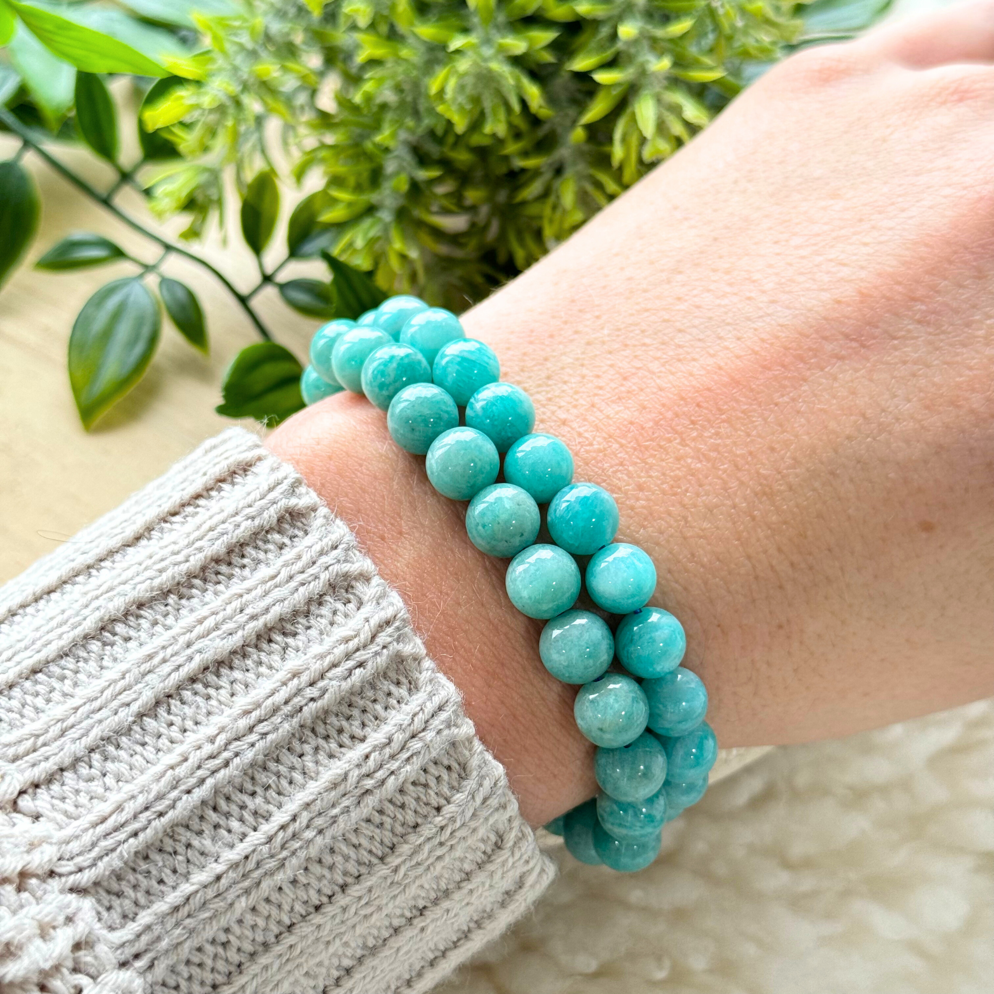 A person's wrist wearing a beaded gemstone bracelet with shades of aqua, teal, and pale green beads, displayed against a neutral background with green foliage in the backdrop.
